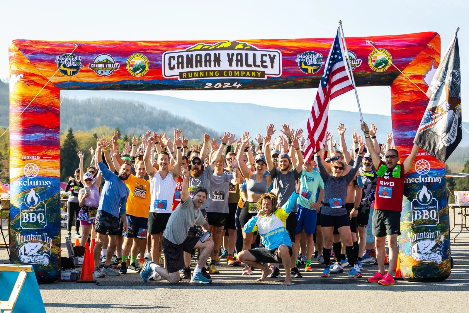 A group of runners cheer and raise their hands under a colorful inflatable arch at the start line of the Canaan Valley Running Company 2024 race, with an American flag waving.