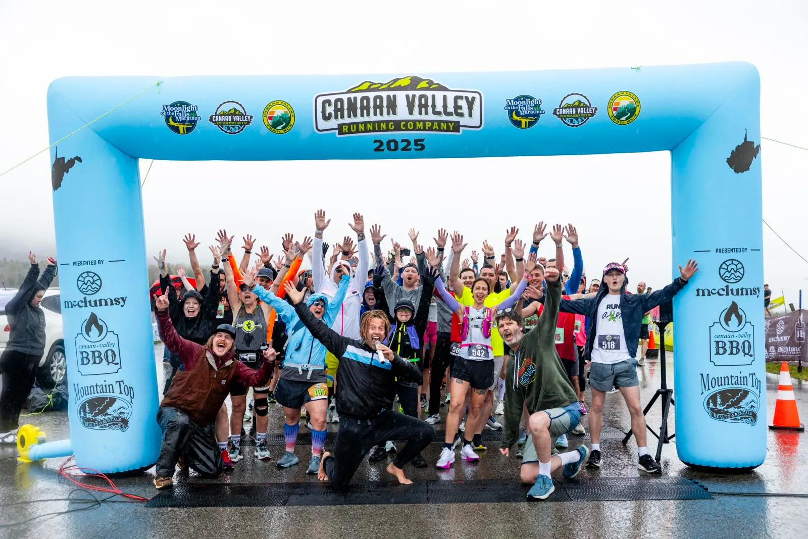 A large group of runners cheer and raise their hands under a blue inflatable archway at the start of the Canaan Valley Running Company 2025 race.