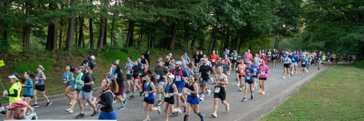 A large group of runners participating in a race on a paved road surrounded by trees.