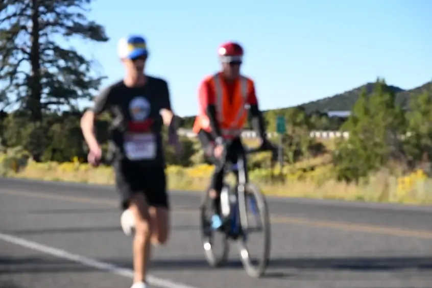 A runner is in motion on a road, with a cyclist in the background, under a clear blue sky with distant hills.