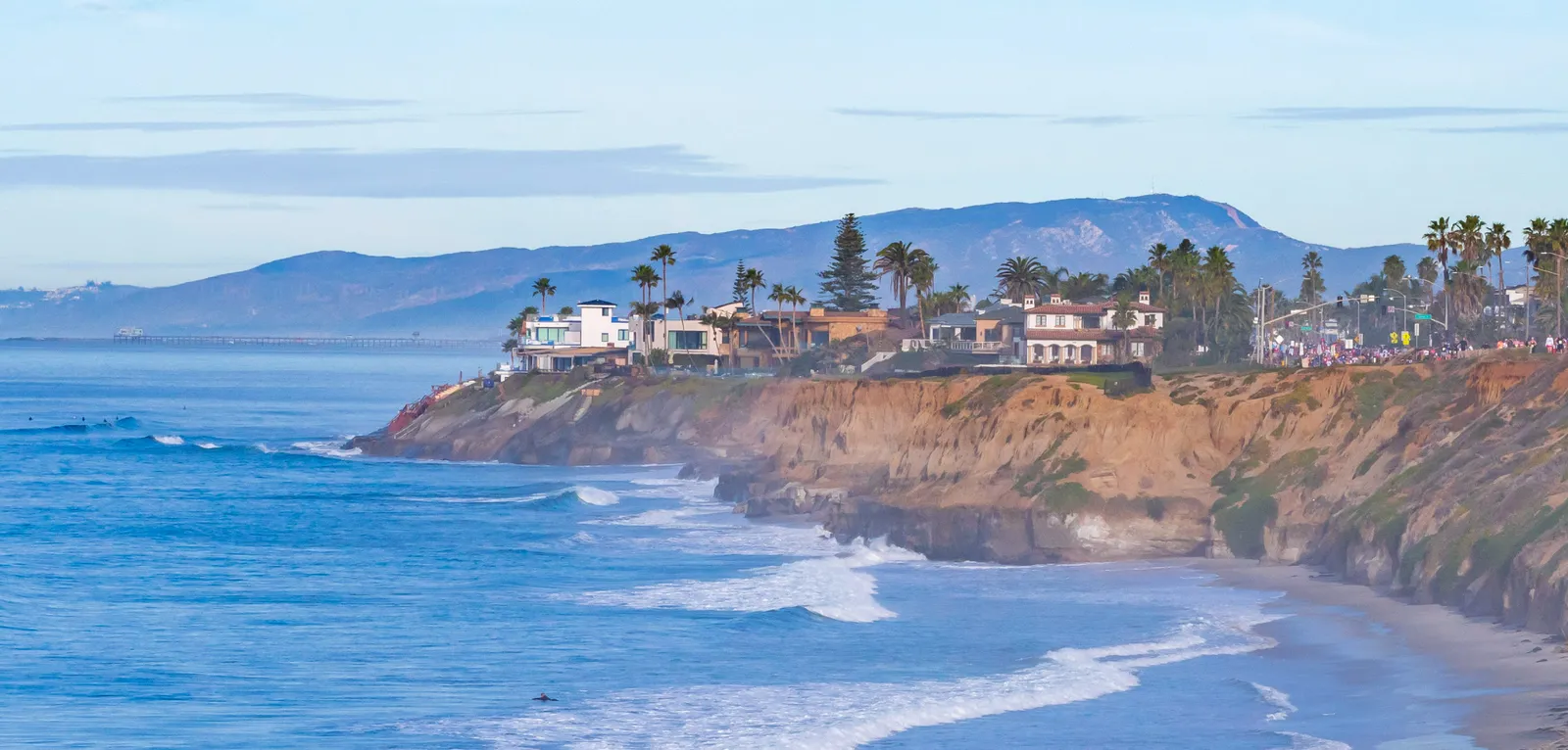 A scenic view of the Carlsbad coastline with ocean waves, cliffs, houses, palm trees, and a distant crowd of runners on the road.
