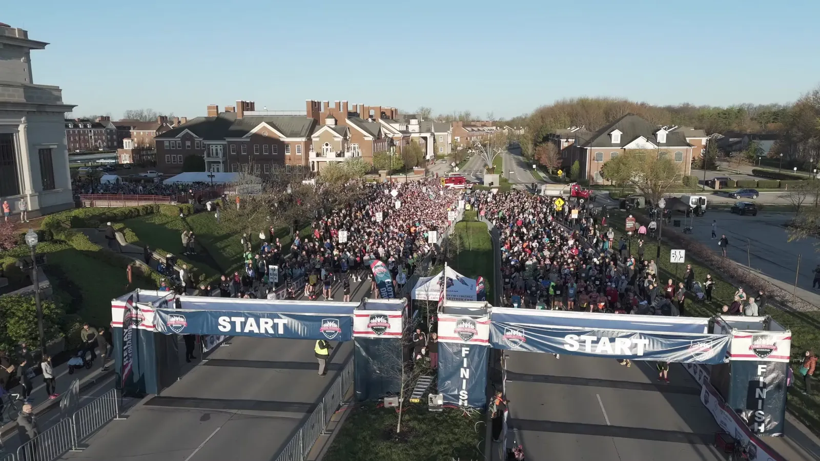 An aerial view of a large crowd of runners gathered at the start line of the Carmel Marathon Weekend, with banners and buildings in the background.