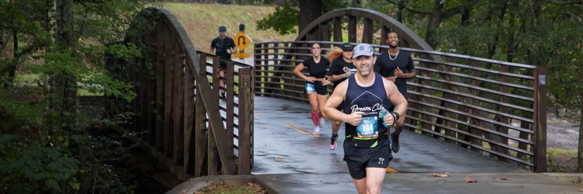 A group of runners crossing a wooden bridge surrounded by trees, with the lead runner smiling at the camera.