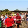 A group of runners, some wearing headbands and hats, are starting a race on a sunny day. The focus is on the runners in the foreground, with more participants and a bridge in the background.