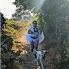A runner wearing a white tutu and a t-shirt with a smiley face runs on a wooded trail with a dog.