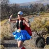 Runner in a superhero costume with a red cape and blue skirt giving a thumbs up on a dirt path with hills in the background.
