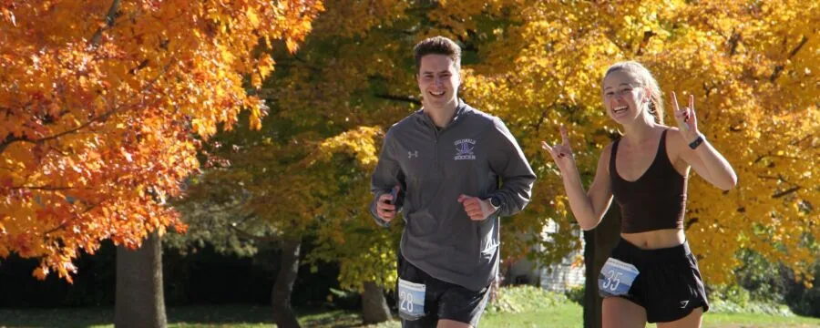 A male and female runner smiling and running with colorful autumn trees in the background.