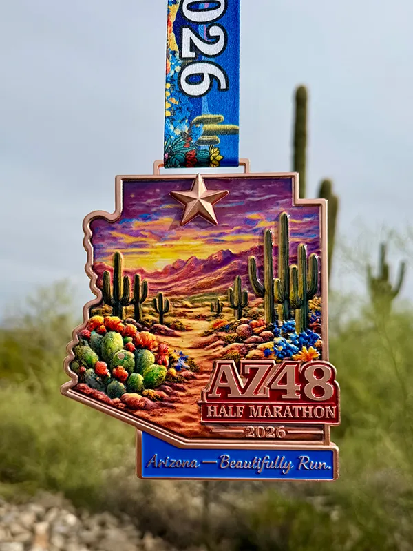 The AZ48 Half Marathon 2026 medal, shaped like Arizona, features a desert landscape with saguaro cacti and a sunset, hanging against a blurred background of desert scenery.