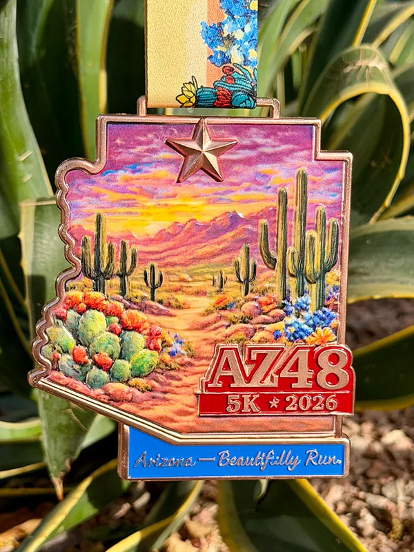 The AZ48 5K 2026 race medal, shaped like Arizona, features a desert landscape with saguaro cacti and a sunset, and the text 'Arizona - Beautifully Run'.