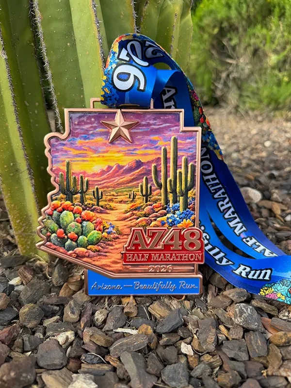 AZ48 Half Marathon 2026 medal with a desert landscape and cactus design, resting on rocks next to a saguaro cactus.