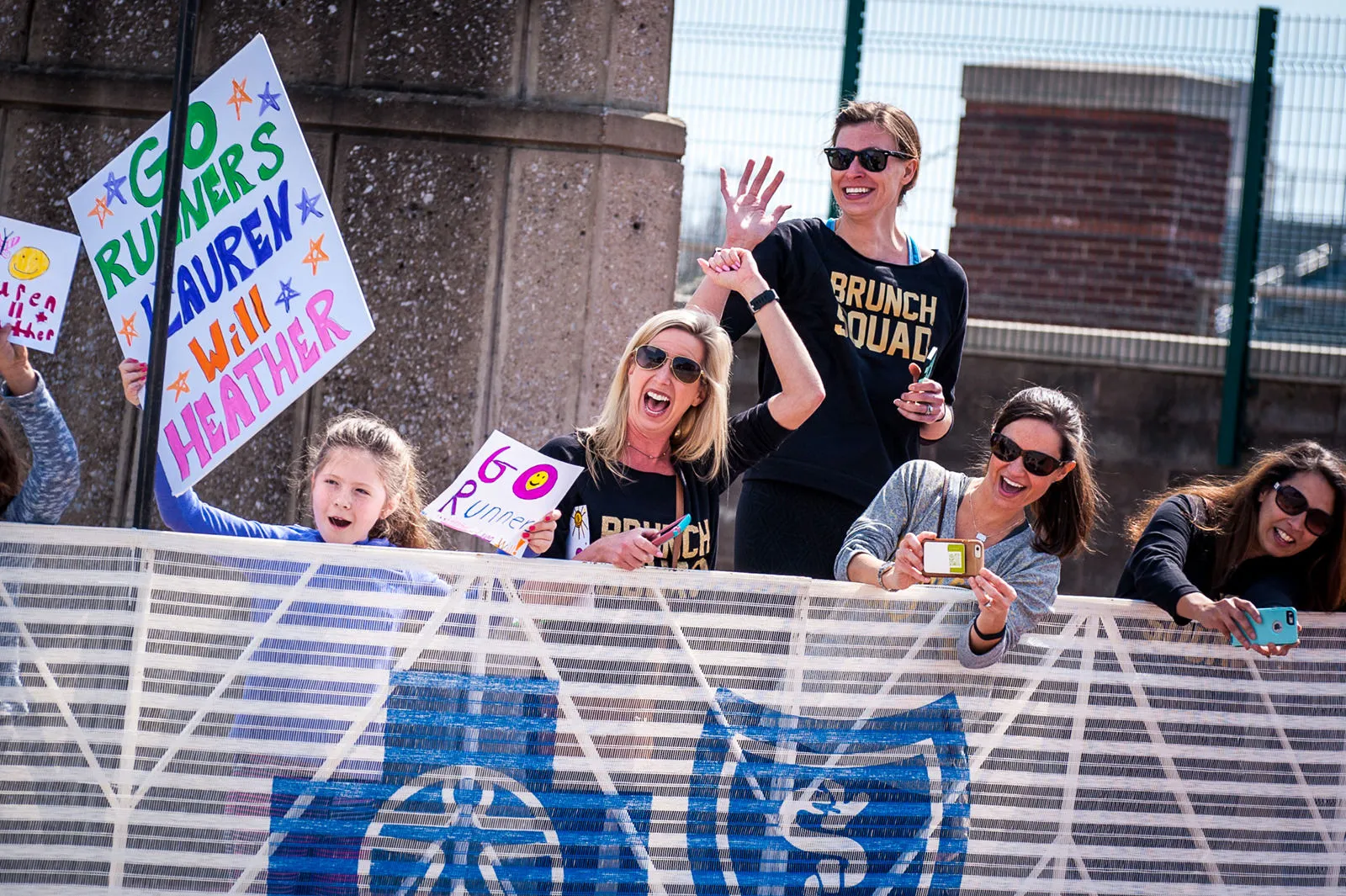 Spectators at a race hold up colorful signs cheering for runners, with one person taking a photo with their phone.