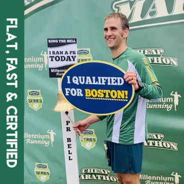 A male runner smiles as he rings a bell and holds a sign that says 'I QUALIFIED FOR BOSTON!' at the finish line of a race.