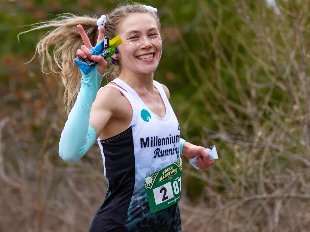 A female runner with glitter on her face smiles and holds up two fingers in a peace sign while running, wearing a bib with the number 287 and 'Millennium Running' branding.