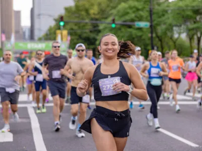 A female runner smiles as she runs in the foreground, with other runners and a 'START' banner visible in the background of the Chicago Spring Half Marathon.