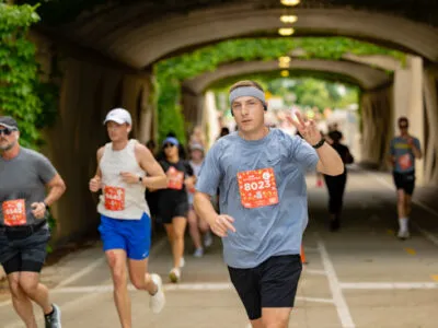 A male runner in the foreground gives a peace sign while running through a tunnel with other runners behind him.