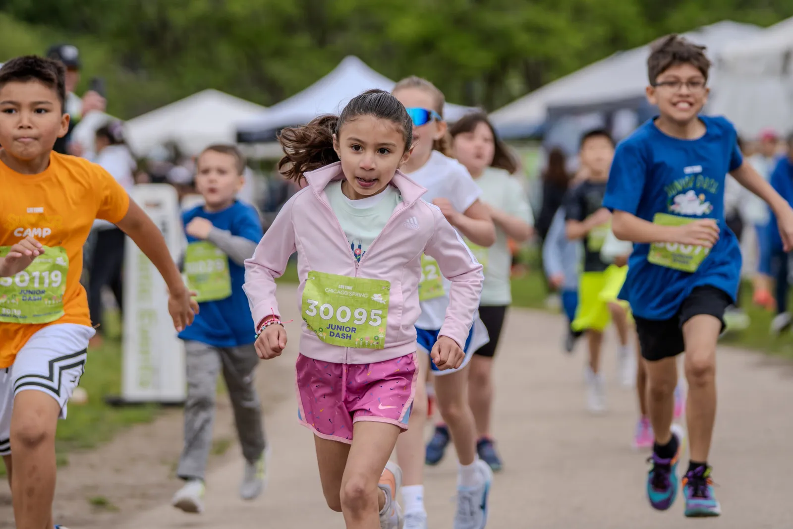 Children running in a race, with bib numbers visible, on a dirt path with tents in the background.