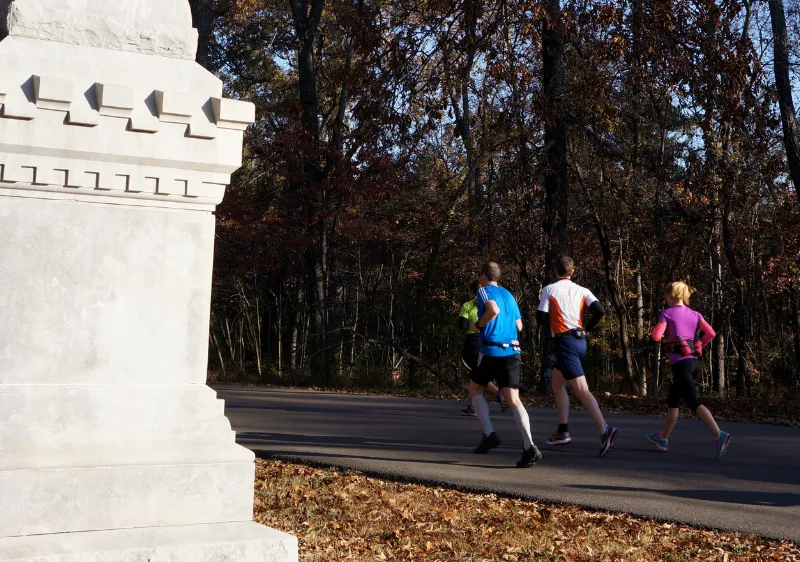 Runners on a paved path through a forest with a stone monument in the foreground.