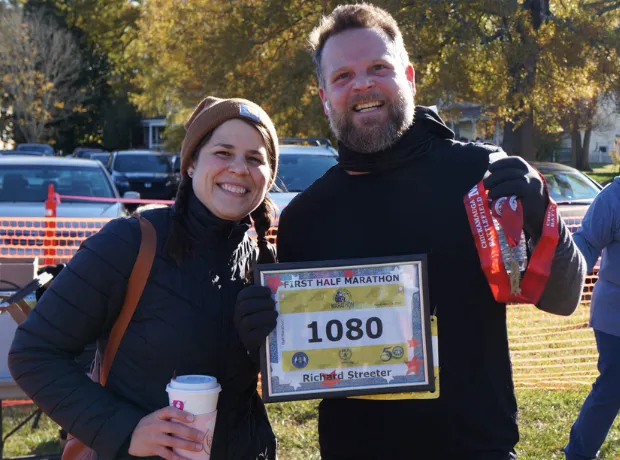 A smiling couple poses for a photo after completing a half marathon. The man holds a framed bib with the number 1080 and the name Richard Streeter, and a race medal around his neck.