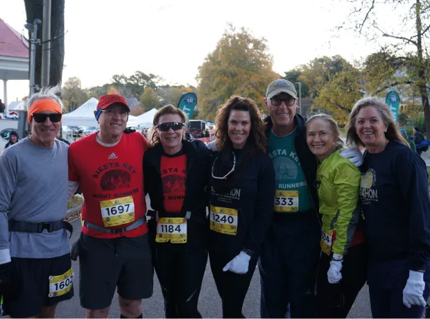 A group of seven runners, smiling and posing for a photo before or after a race. They are wearing race bibs and some have race-branded shirts.