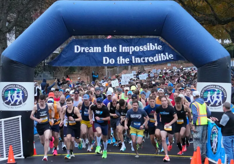 Runners surge forward under a blue inflatable arch at the start of a race, with a banner reading 'Dream the Impossible Do the Incredible' overhead.