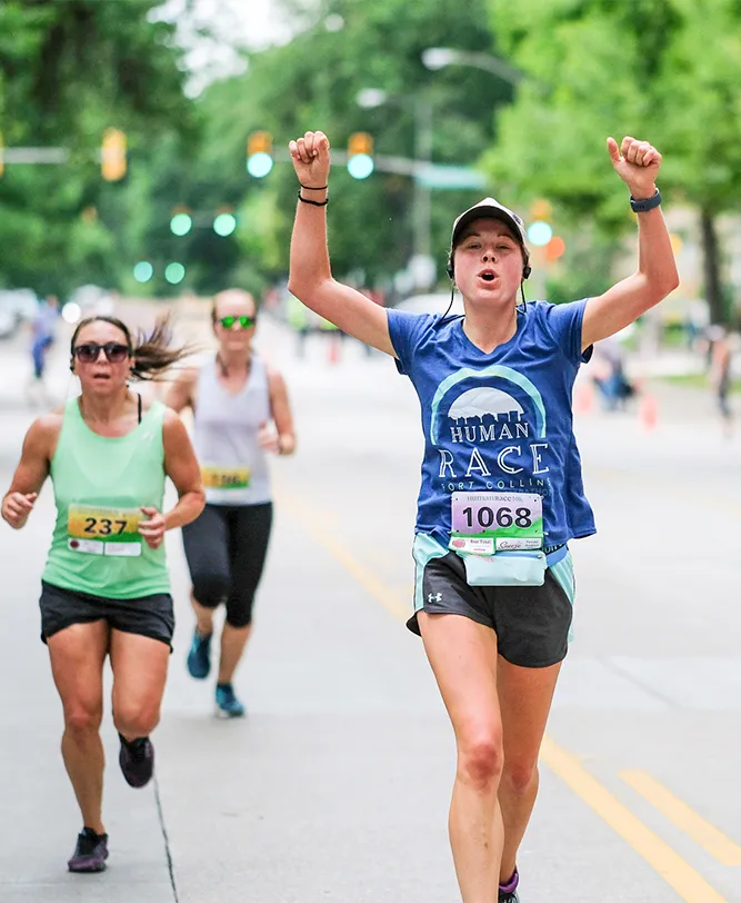 A female runner with her arms raised in celebration crosses the finish line of a road race.