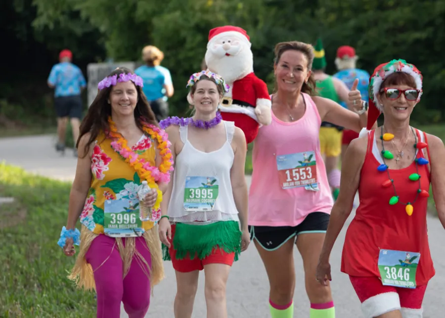 Four women in festive Christmas-themed costumes, including leis and Santa hats, running a race and smiling at the camera.