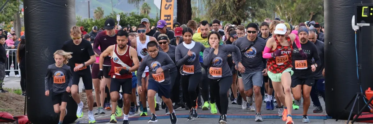 A large group of runners are starting a race, with some in the foreground and others further back. The start line arch is visible, and the runners are all looking forward.