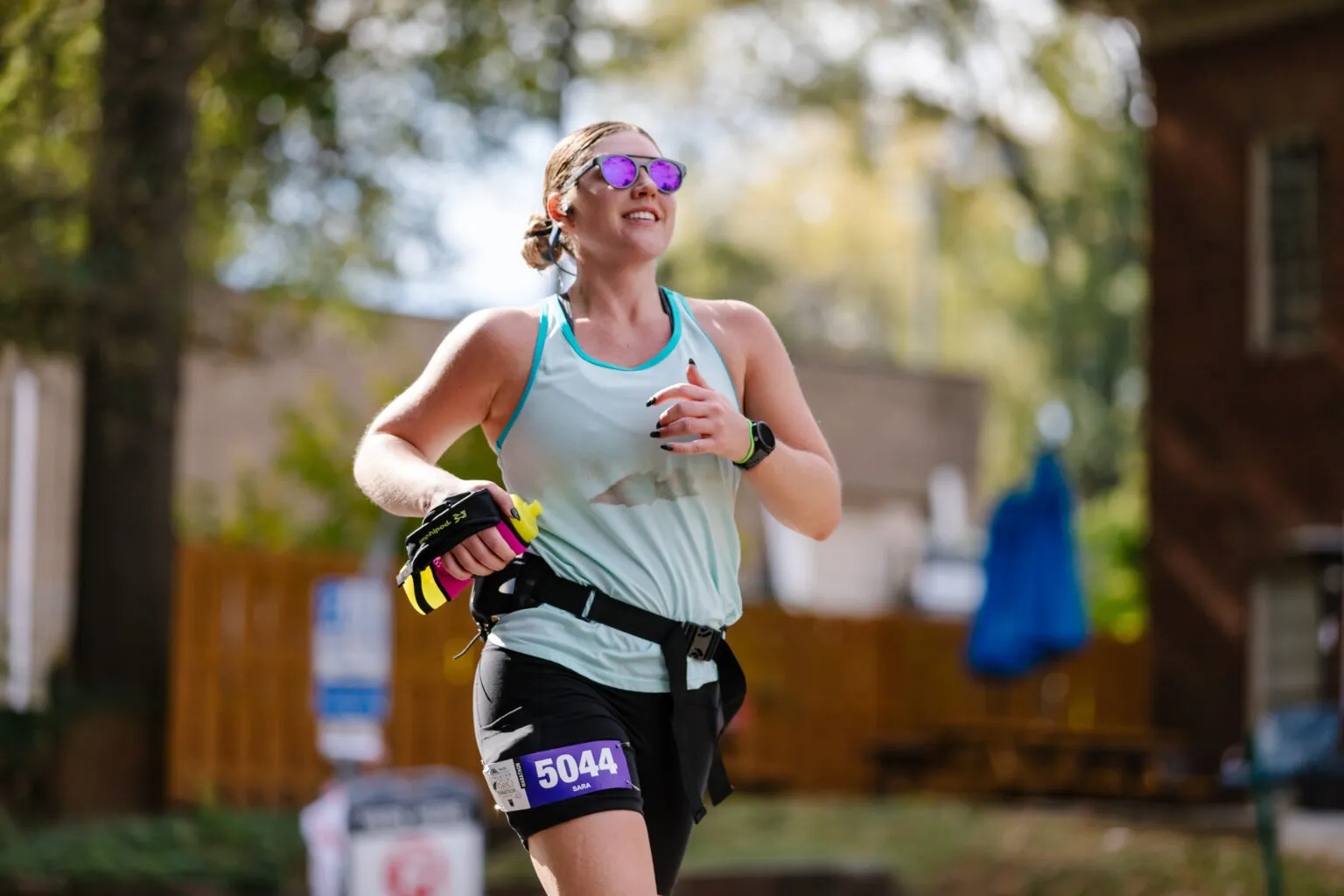 A female runner smiles as she runs, wearing sunglasses and a race bib with the number 5044.