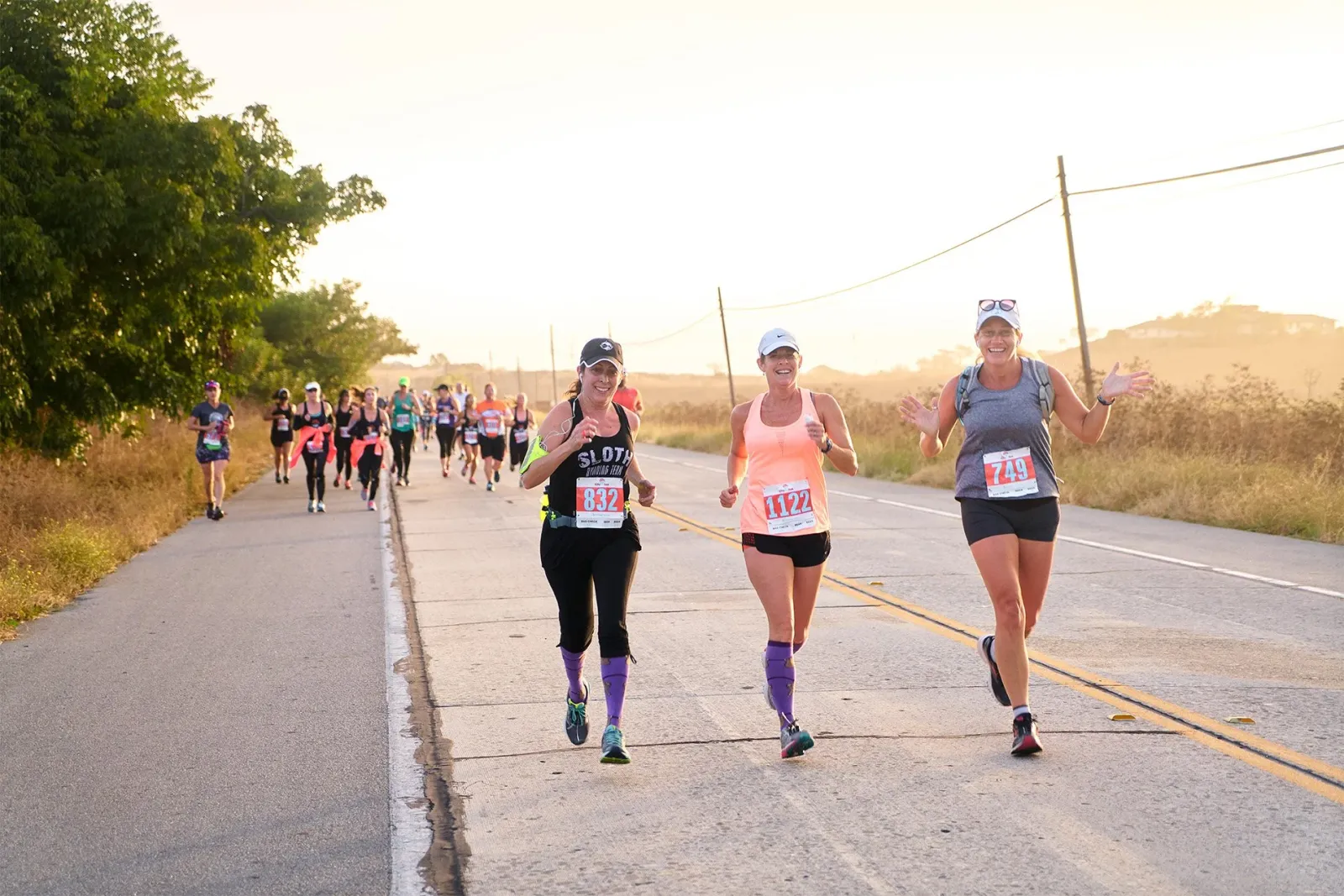 Three women running a race on a road with other runners in the background, with the sun setting.