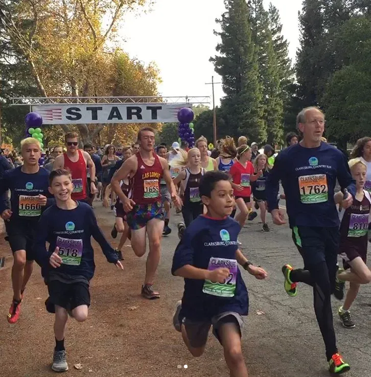 A group of runners, including children and adults, are starting a race under a 'START' banner. Many are wearing matching race shirts.