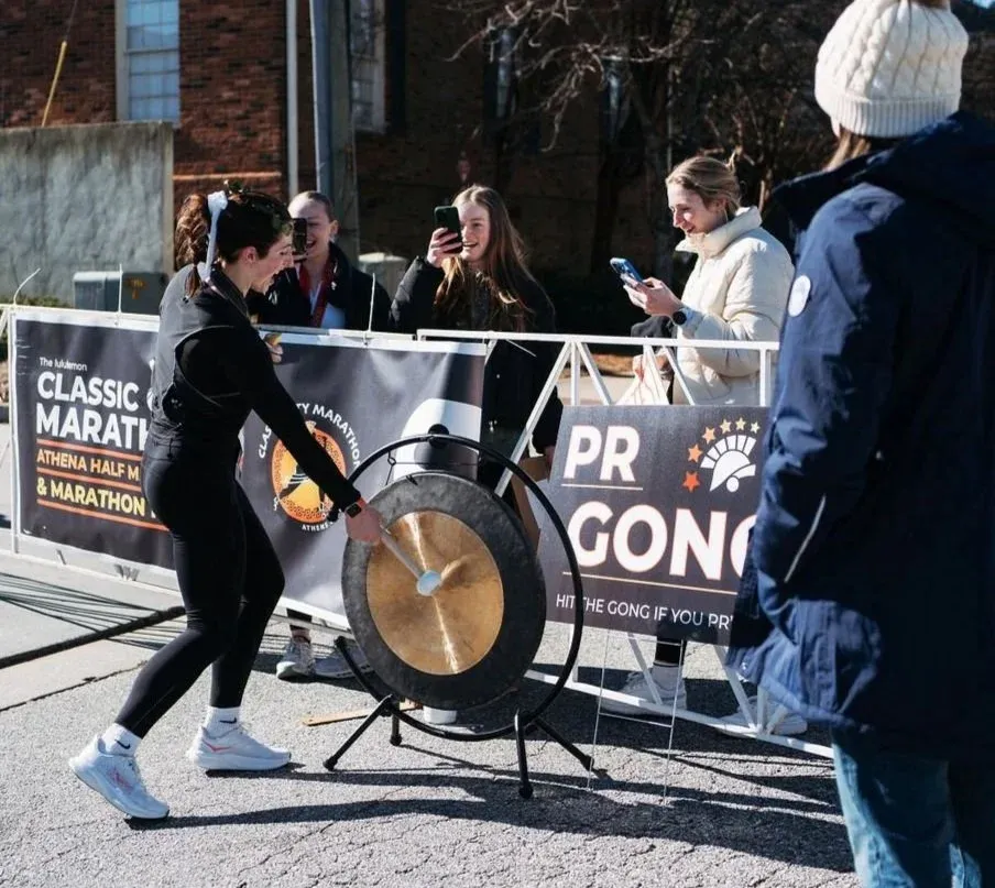 A female runner in black athletic wear strikes a large gong with a mallet, with spectators and race banners in the background.