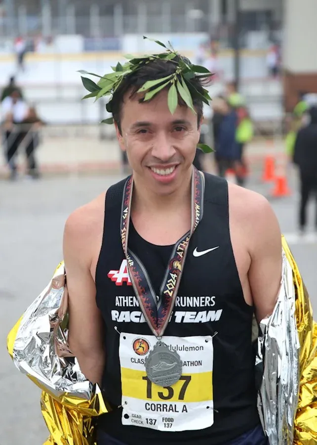 A smiling runner wearing a laurel wreath and a medal, wrapped in a silver thermal blanket, after finishing a race.