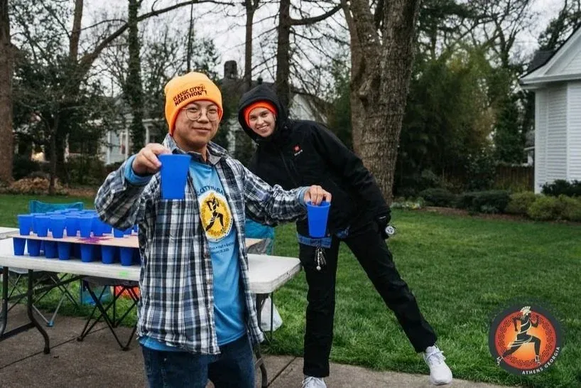 Two people, possibly volunteers, stand behind a table with many blue cups, holding cups themselves and smiling.