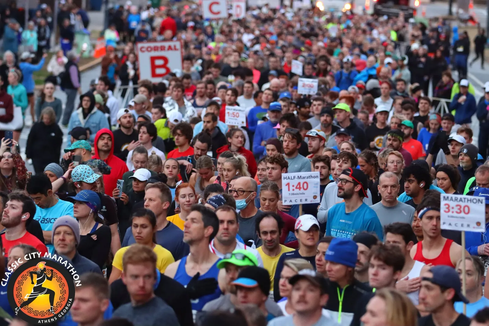 A large crowd of runners gathered at the start line of a marathon, with signs indicating race corrals and pacers visible.