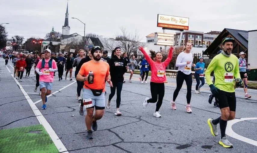 A group of runners starting a race on an urban street, with one runner in the foreground wearing a bright green long-sleeved shirt and another in a pink shirt with arms raised in excitement.