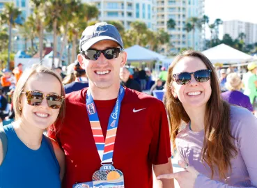 Three smiling people, two women and one man wearing a race medal, pose for a photo at an outdoor event.