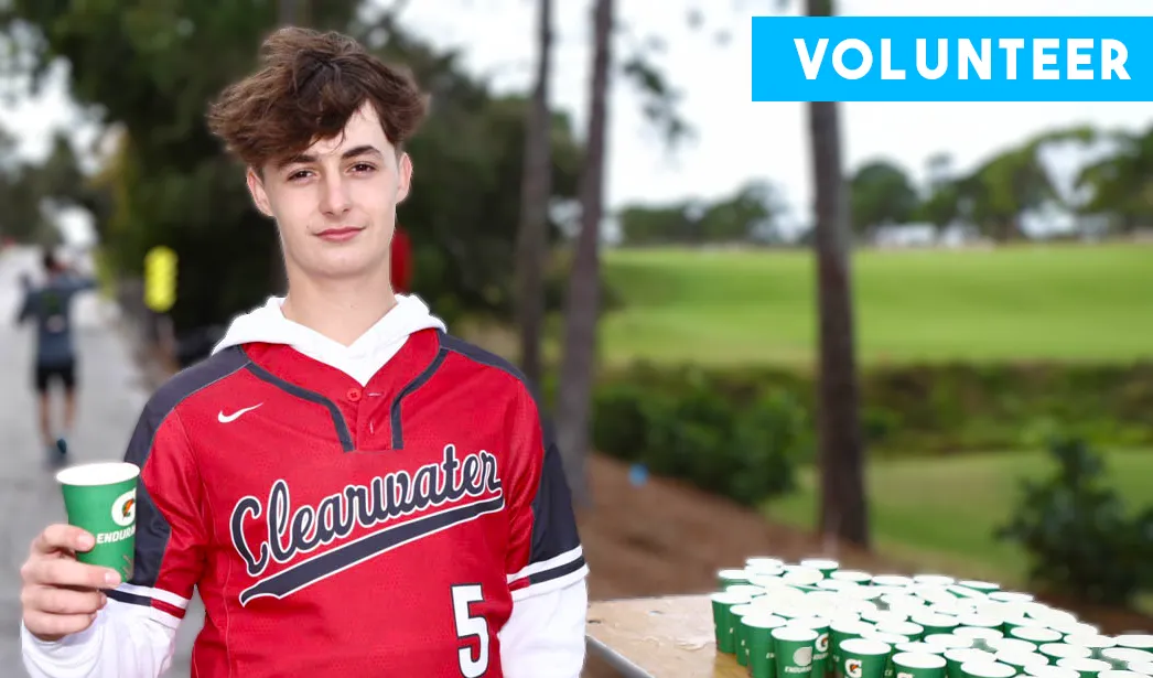 A young person wearing a 'Clearwater' jersey and holding a Gatorade cup, with a 'VOLUNTEER' banner at the top and a blurred background of a race course.