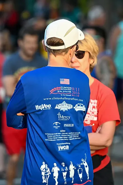 A runner in a blue long-sleeved shirt with race details and sponsor logos, standing with another person at what appears to be the start line of the Clinton Half Marathon.