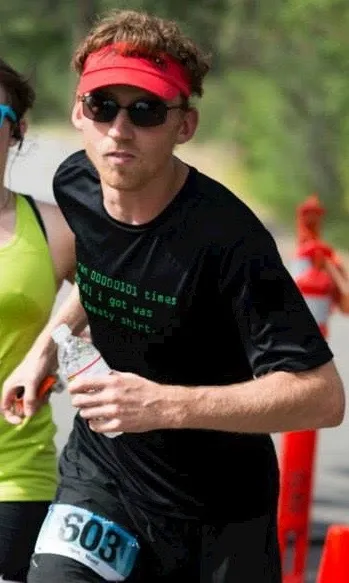A male runner wearing a red visor, sunglasses, and a black t-shirt with binary code text, running with a water bottle and bib number 603.