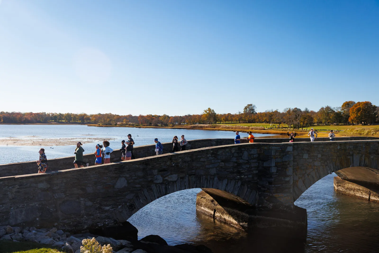 Runners crossing a stone bridge over water with autumn foliage in the background.