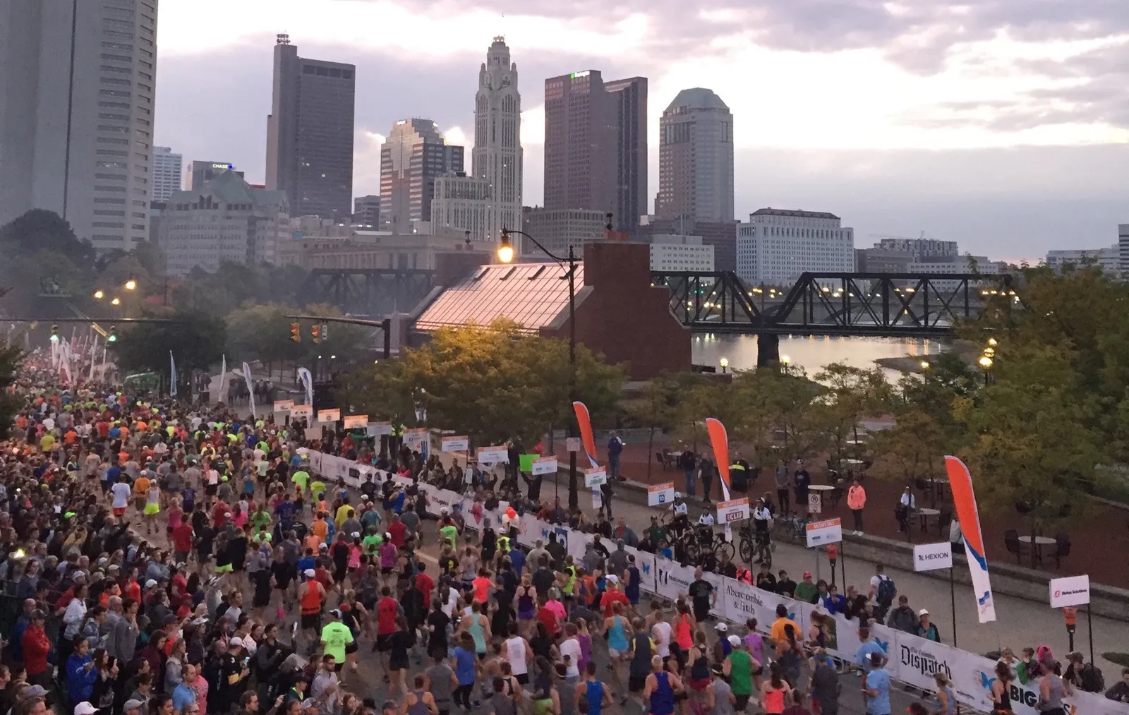 A large crowd of runners on a city street with bridges and skyscrapers in the background during a marathon.