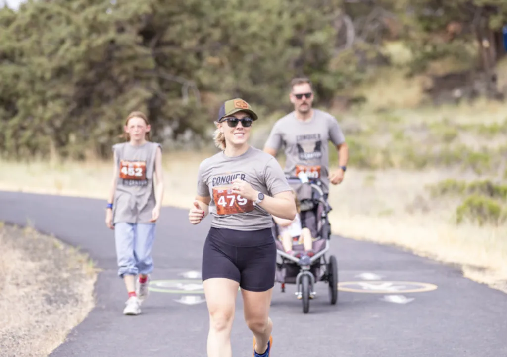 A woman in the foreground runs with a smile, wearing a race bib with the number 479. Behind her, a young person runs with bib 452, and a man pushes a stroller, also wearing race attire.