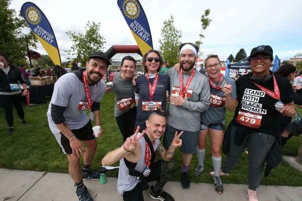 A group of six smiling runners, some wearing medals, pose for a photo after a race, with Rotary banners in the background.