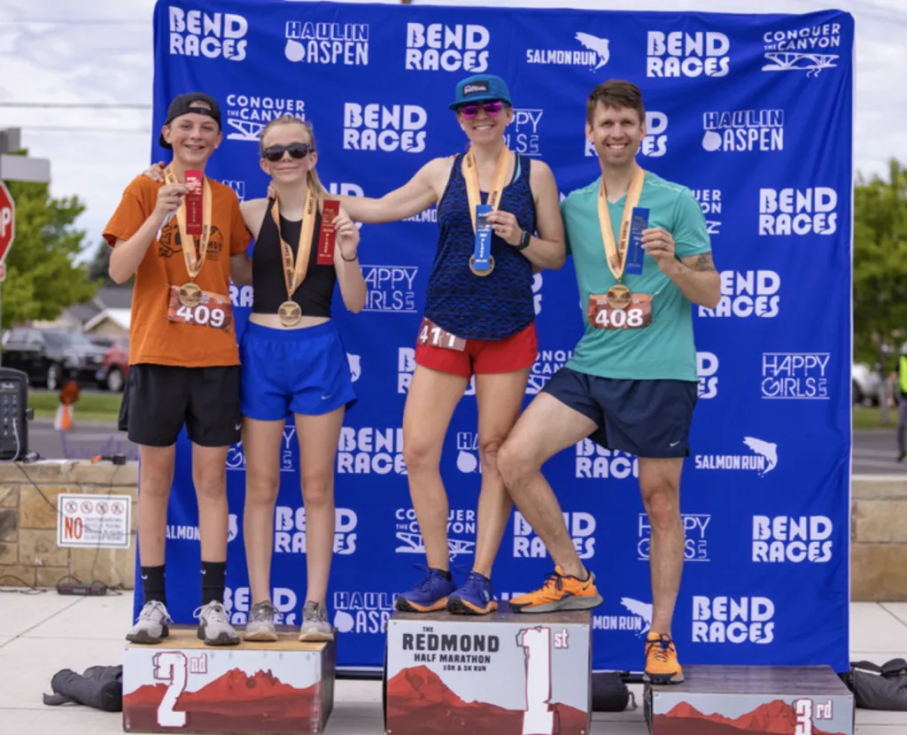 Four runners, two adults and two children, stand on a podium holding medals and ribbons, with a blue banner displaying race sponsor logos in the background.