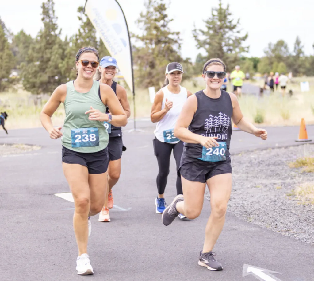 Three women running a race, wearing bib numbers and athletic clothing, with trees and a banner in the background.