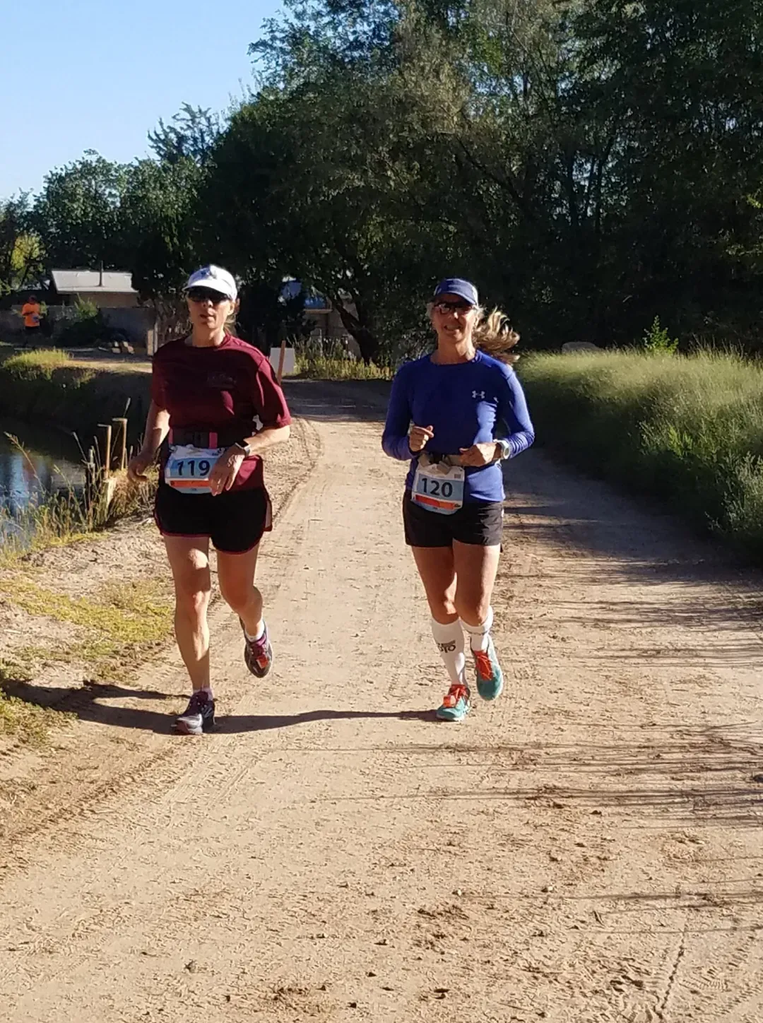 Two female runners on a dirt path, one in a maroon shirt and the other in a blue shirt, both wearing race bibs.