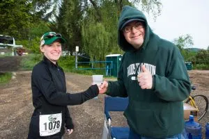 A female runner with bib number 1078 shakes hands with a male runner wearing a green hooded sweatshirt and giving a thumbs up.