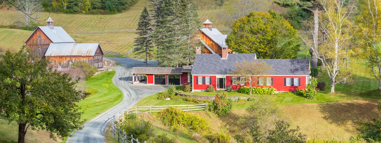 A scenic view of a rural Vermont farm with red and wooden barns, a winding gravel road, and surrounding trees in autumn colors.