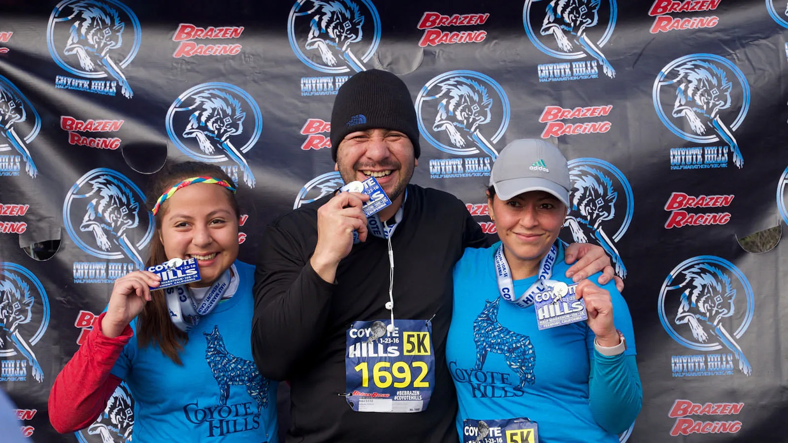 Three smiling people, two women and one man, pose for a photo with race medals around their necks and bibs visible. They are standing in front of a banner with race logos and branding.