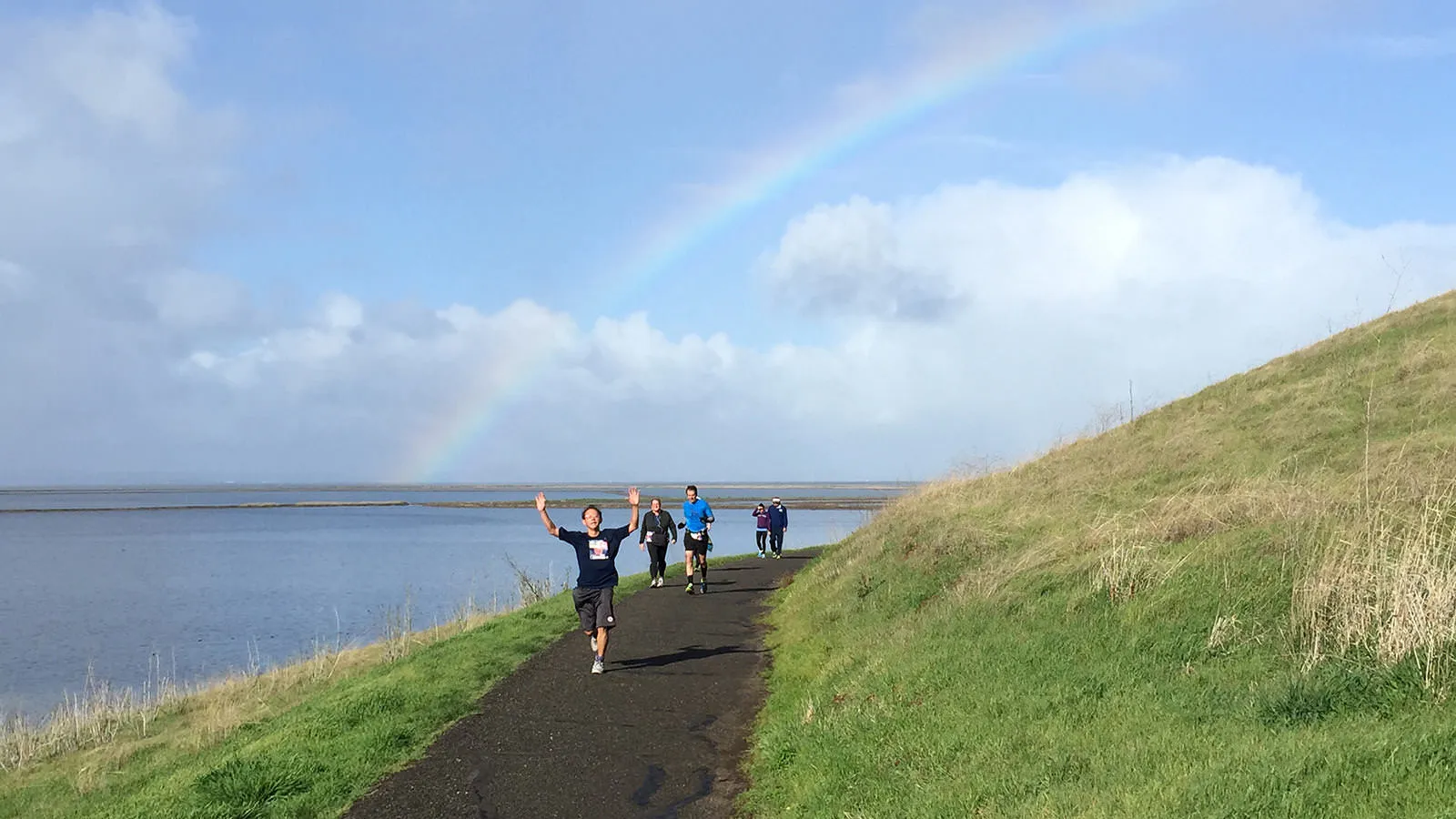 A runner with arms raised in celebration runs on a path next to a body of water, with a rainbow arching across the sky.